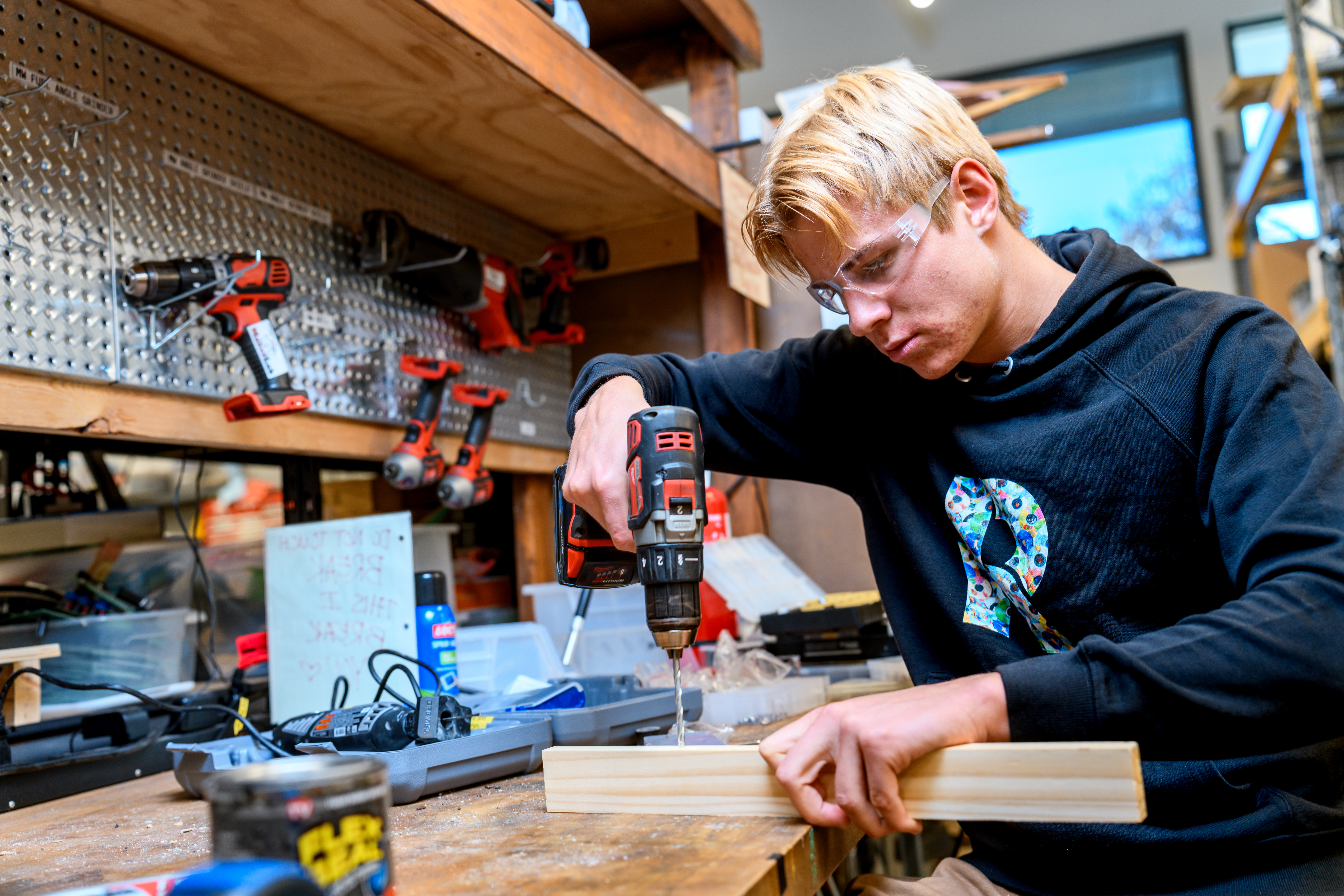 A student wearing safety goggles uses a drill and woodworking equipment.